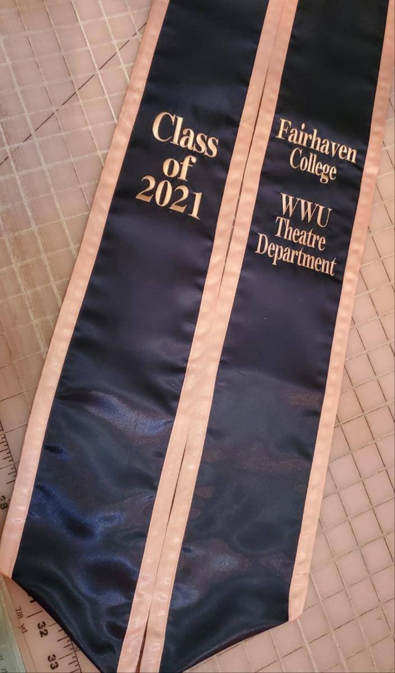 May include: A black graduation sash with peach colored trim. The sash has gold embroidery that reads "Class of 2021", "Fairhaven College", and "WWU Theatre Department".