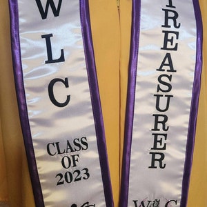 May include: White and purple graduation sash with the text "WLC Class of 2023" and "Treasurer" on it. The sash also has a logo for the Women's Law Caucus.