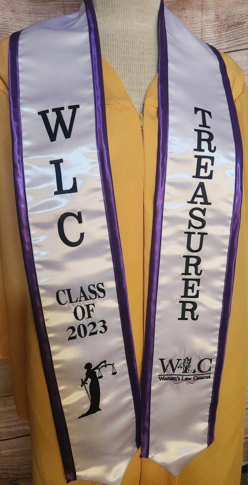 May include: White and purple graduation sash with the text "WLC Class of 2023" and "Treasurer" on it. The sash also has a logo for the Women's Law Caucus.