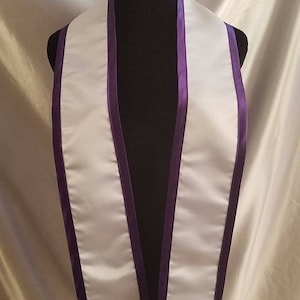 May include: White satin graduation stole with purple trim. The stole is worn around the neck and hangs down the front of the body.