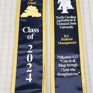 May include: A navy blue graduation stole with gold trim. The stole features the North Carolina Agricultural & Technical State University logo, the text "Class of 2024", and a Bible verse from Philippians 4:13. The stole also includes the text "B.S. Business Management".
