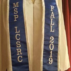May include: A white graduation gown with a blue and white sash. The sash has the text "MSP LCSR" on one side and "FALL 2019" on the other side.