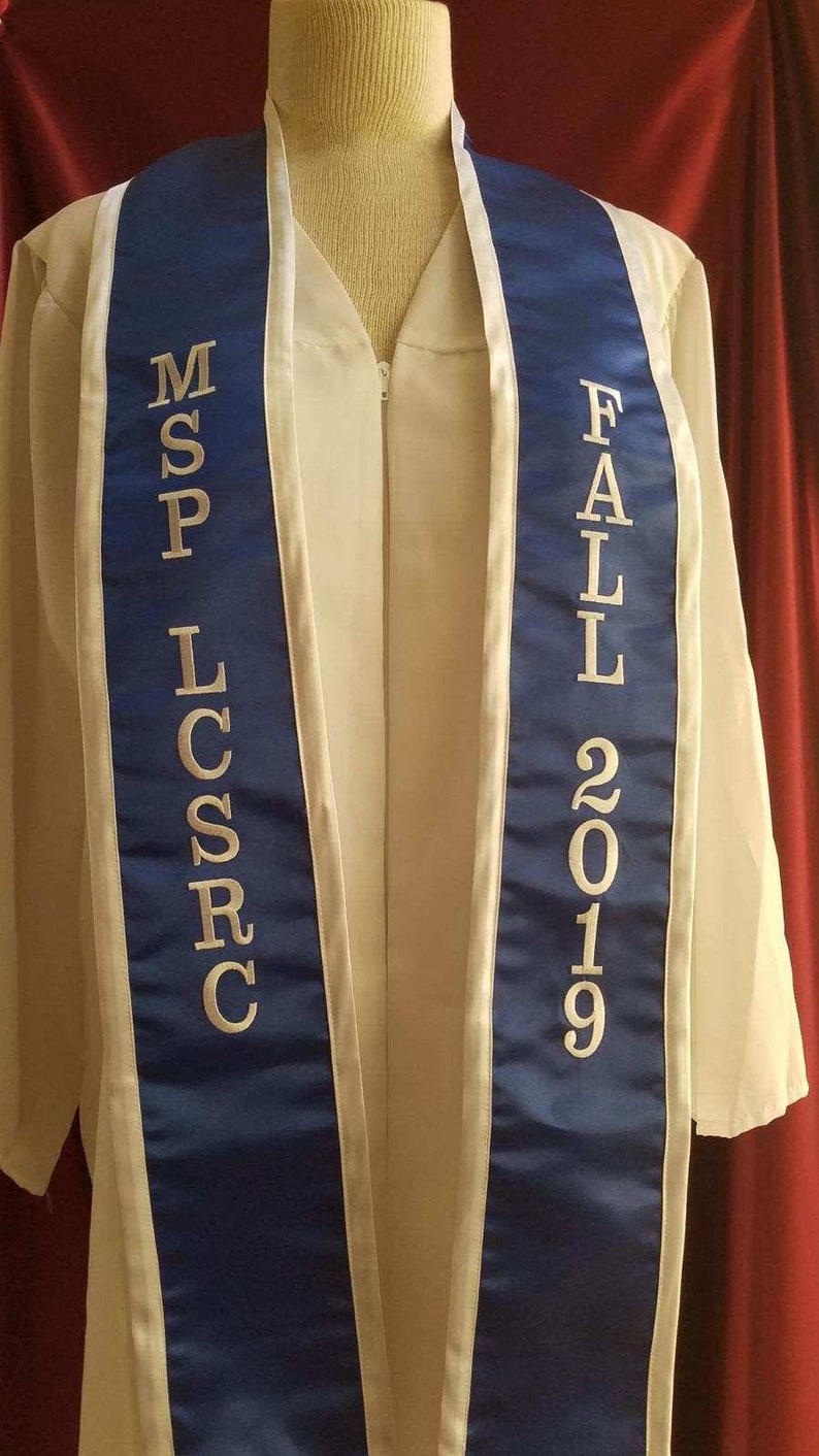 May include: A white graduation gown with a blue and white sash. The sash has the text "MSP LCSR" on one side and "FALL 2019" on the other side.