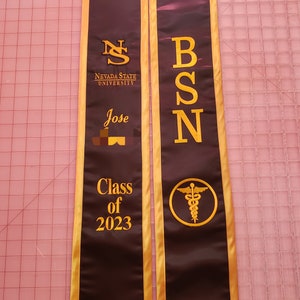 May include: A brown graduation stole with gold trim. The stole has the text "Nevada State University" and "Class of 2023" on one side. The other side has the text "BSN" and a caduceus symbol.
