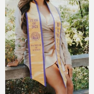 May include: A woman wearing a beige dress and a gold and purple graduation sash. The sash says "Class of 2021" and "Bachelor of Science".