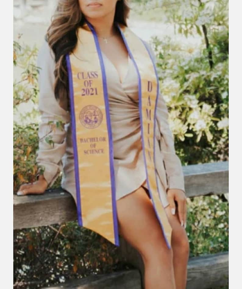 May include: A woman wearing a beige dress and a gold and purple graduation sash. The sash says "Class of 2021" and "Bachelor of Science".