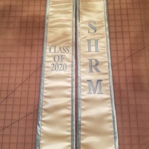 Może przedstawiać: Para szarf graduation, jedna z napisem "CLASS OF 2020", a druga z napisem "SHRM". Szarfy są jasnozłote z niebieskim wykończeniem.