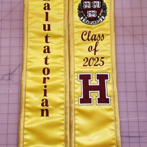 May include: Yellow graduation sash with maroon text that reads "Salutatorian" on the left side and "Class of 2025" and a large maroon "H" on the right side. The sash also features a maroon Harvard University crest.