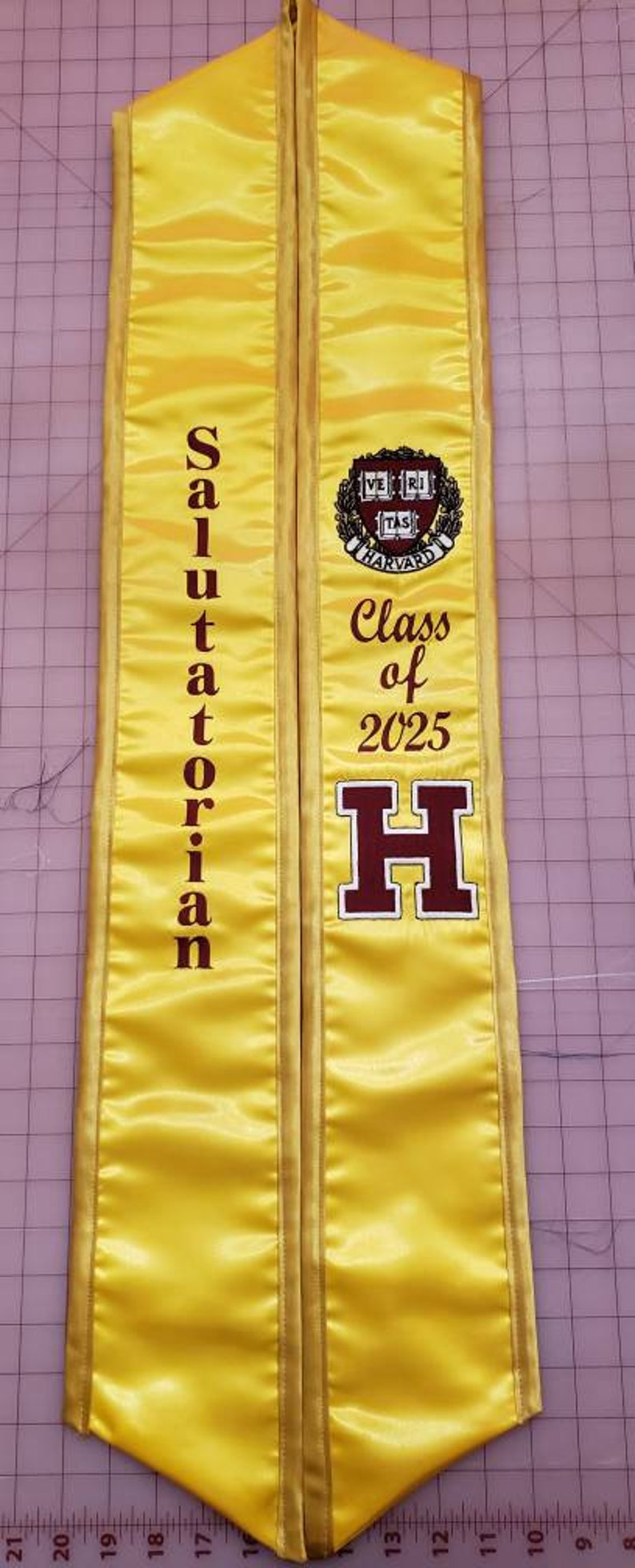 May include: Yellow graduation sash with maroon text that reads "Salutatorian" on the left side and "Class of 2025" and a large maroon "H" on the right side. The sash also features a maroon Harvard University crest.