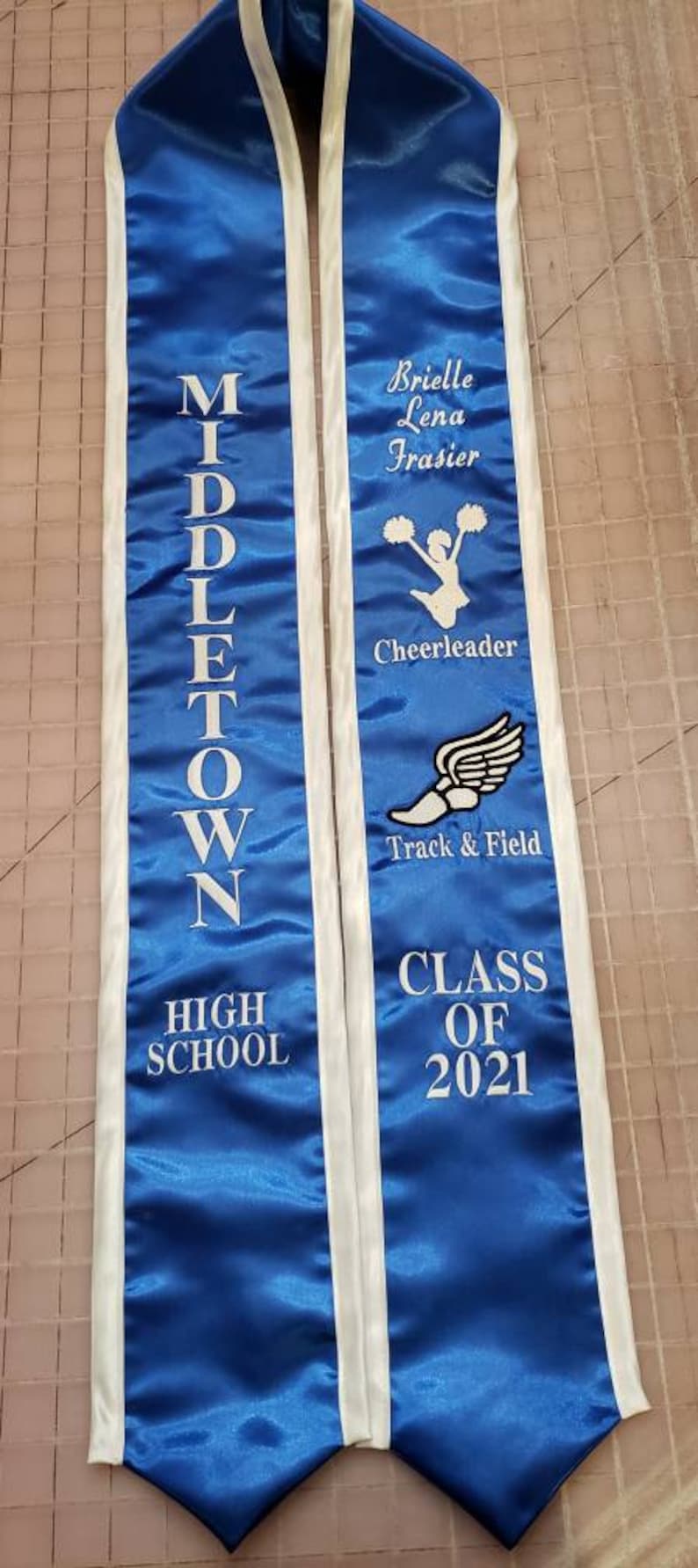 May include: Blue and white graduation sash with white text that reads "Middletown High School" on the left side and "Brielle Lena Frasier Cheerleader Track & Field Class of 2021" on the right side.