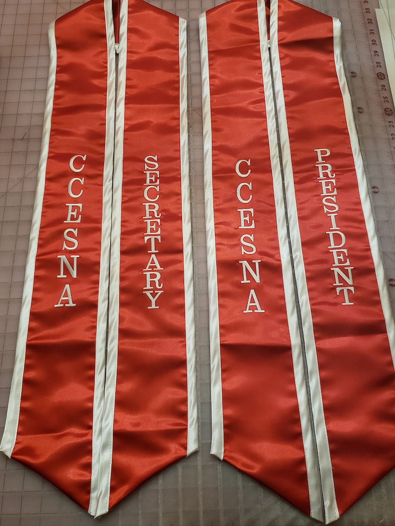 May include: Two red satin graduation sashes with white piping. The sashes have the words "CESNA SECRETARY" and "CESNA PRESIDENT" embroidered in white.
