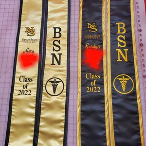 May include: Two gold and black graduation sashes with the text "Nevada State College" and the degree "BSN" on each. The sashes also have the text "Class of 2022" and a caduceus symbol. The names "Alexa" and "Rosalyn" are written on the sashes.
