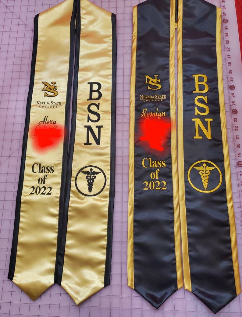 May include: Two gold and black graduation sashes with the text "Nevada State College" and the degree "BSN" on each. The sashes also have the text "Class of 2022" and a caduceus symbol. The names "Alexa" and "Rosalyn" are written on the sashes.