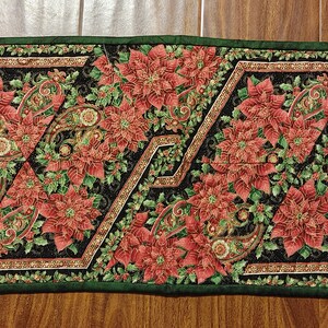 May include: A green and black table runner with a repeating pattern of red poinsettia flowers and paisley designs. The runner has a scalloped edge.