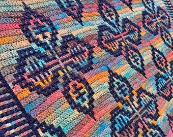 Op de afbeelding: Een kleurrijk gehaakt deken met een geometrisch patroon in tinten blauw, roze, oranje en bruin. Het deken heeft een donkerblauwe rand.