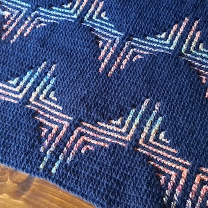 Könnte beinhalten: Eine marineblaue Häkeldecke mit einem geometrischen Muster in Rosa-, Orange- und Blautönen.