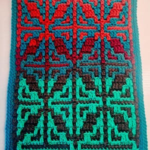 May include: A handmade crochet piece with a geometric pattern in shades of red, teal, brown, and green. The design features repeating cross-like shapes and is bordered by a teal edge. The texture of the yarn is visible.