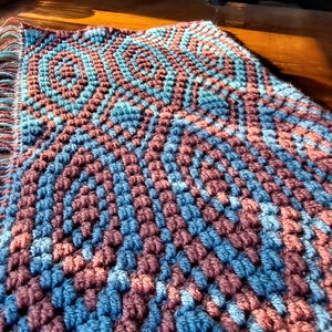 May include: Handmade crochet scarf in a geometric pattern. The scarf features alternating rows of blue and burgundy yarn, with a fringe of matching colors. The scarf is displayed on a wooden surface.