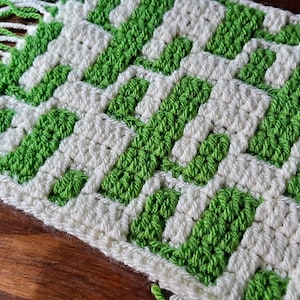 May include: A white and green crocheted scarf with fringe on the ends. The scarf has a geometric pattern.