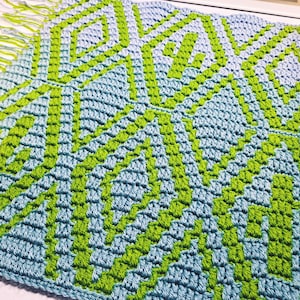 May include: A handmade crochet blanket with a geometric pattern in light blue and green. The design features diamond and zig-zag shapes. The blanket has a fringe edge.