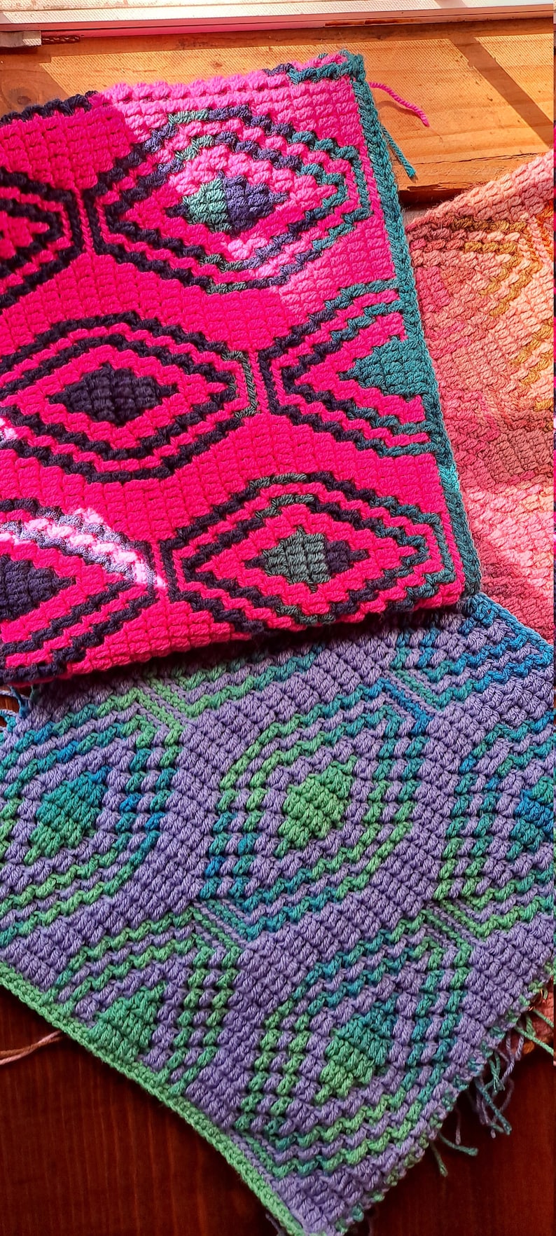Może przedstawiać: Zbliżenie na trzy kolorowe, teksturowane elementy szydełkowe. G&oacute;rny element jest w kolorze gorącego r&oacute;żu z czarnymi i turkusowymi geometrycznymi wzorami. Poniżej znajduje się fioletowy, zielony i niebieski element z podobnymi wzorami. Częściowo widoczny jest element w kolorze brzoskwiniowym.