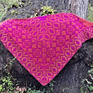 May include: A pink and orange crocheted shawl with a geometric pattern. The shawl is draped over a tree trunk.