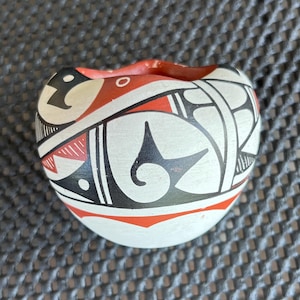 May include: A small, white ceramic pot with a red and black geometric design. The pot is shaped like a heart and has a small opening at the top.