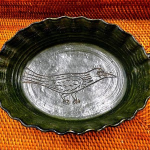 May include: Oval-shaped ceramic serving dish with a dark green scalloped rim and handles. The interior features a silver-colored surface with a detailed bird illustration. A decorative piece for serving food or displaying.