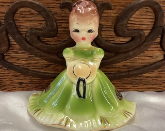 May include: A ceramic figurine of a girl wearing a green dress and holding a straw hat. The figurine has a painted face with closed eyes and red lips.