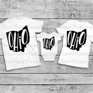 May include: Three white t-shirts with a black outline of the state of Ohio and the word "Ohio" written inside. The shirts are in different sizes, with the largest shirt on the left, the middle shirt in the center, and the smallest shirt on the right.