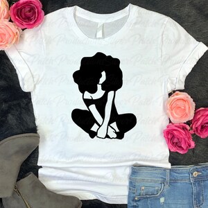 May include: White t-shirt with a black silhouette of a woman with an afro sitting with her legs crossed.