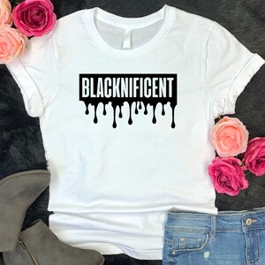 May include: White t-shirt with a black graphic that says "BLACKNIFICENT" in dripping letters.