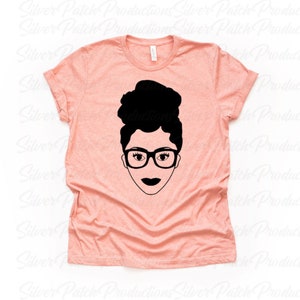May include: A peach colored t-shirt with a black and white illustration of a woman with glasses and her hair in a bun.