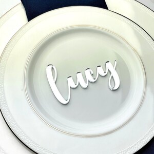 May include: A white plate with a gold rim and a silver acrylic place card with the name "Lucy" on it. The plate is on top of a larger white plate with a black rim. A navy blue napkin is underneath the plates.