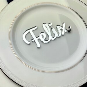 May include: Silver mirror place card with the name "Felix" written in cursive.
