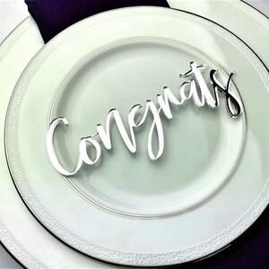 May include: Silver 'Congrats' sign on a white plate with a black and gold rim. The plate is on top of a second white plate with a black rim. A purple napkin is underneath the plates.