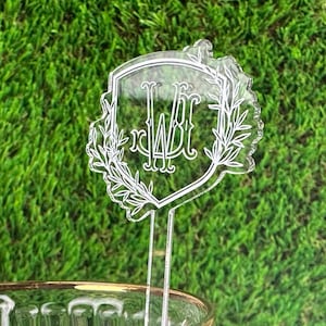 Puede incluir: Agitador de bebidas de acr&iacute;lico transparente con un dise&ntilde;o en forma de escudo que presenta un monograma "WBT" rodeado de ramas con hojas.