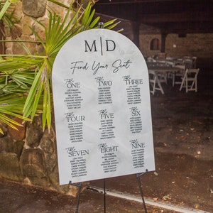 May include: A white arch-shaped seating chart with black text. The chart is labeled "M/D Find Your Seat" and lists tables one through nine with guest names.