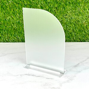 May include: A white frosted acrylic sign holder with a rounded top and a silver base. The sign holder is standing on a white marble surface.