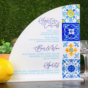 May include: A white sign with a blue and yellow tile pattern. The sign lists signature cocktails, beer and wine, and spirits. The text includes "The Lulu", "The Ringo", "Blue Moon", "Coors Light", "Miller", "Heineken", "KJ Chardonnay", "Decoy Merlot", "Hendricks Gin", "Casamigos Reposado", "Titos Vodka", "Crown Royal", and "Bulleit Rye Whiskey".