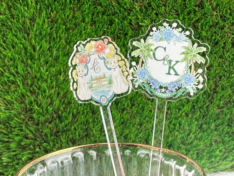 Puede incluir: Dos toppers de pastel de acr&iacute;lico con dise&ntilde;os florales. Un topper presenta una casa blanca con un techo azul y dos gatos blancos. El otro topper presenta un escudo verde con las letras "C & K" y el texto "Est. 2022".
