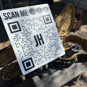 May include: A white sign with a black QR code and the text "SCAN ME" with icons for Instagram, Cash App, and a document. The text "JH" is in the center of the QR code. The sign is propped up on a black guitar case.