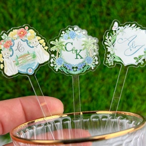 May include: Three clear acrylic drink stirrers with floral designs and personalised text. The first stirrer has a floral design with two cats and the text "L & S". The second stirrer has a floral design with palm trees and the text "C & K". The third stirrer has a floral design with the text "A & J".