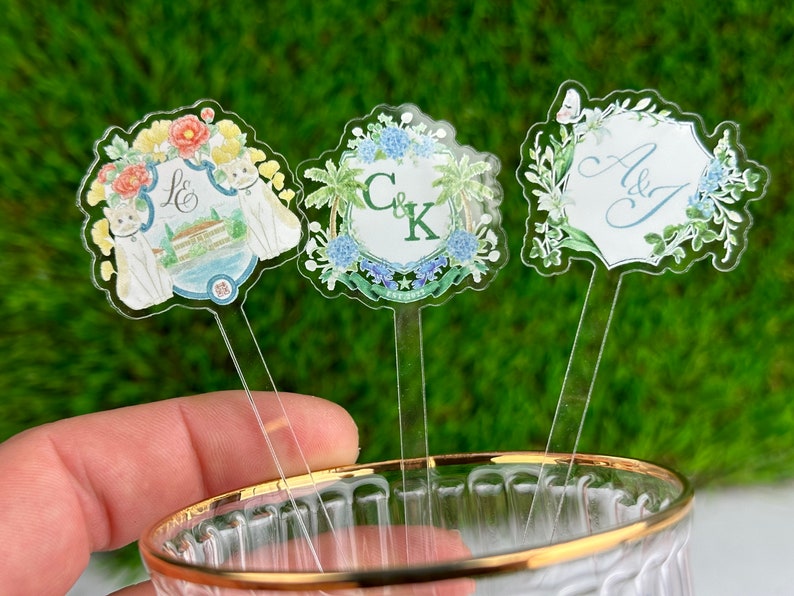 May include: Three clear acrylic drink stirrers with floral designs and personalised text. The first stirrer has a floral design with two cats and the text "L & S". The second stirrer has a floral design with palm trees and the text "C & K". The third stirrer has a floral design with the text "A & J".