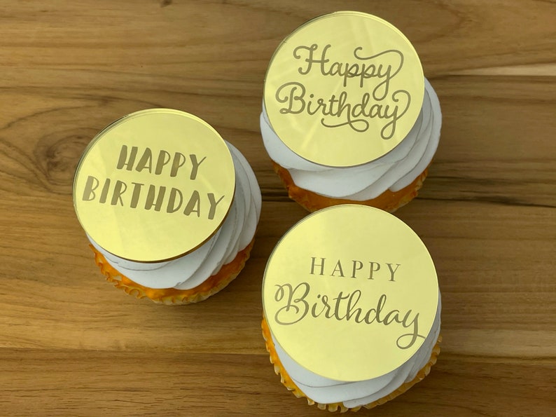 Może przedstawiać: Trzy złote ozdoby do babeczek z napisem "Happy Birthday" w r&oacute;żnych czcionkach. Ozdoby znajdują się na białych babeczkach.