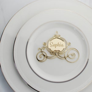 Op de afbeelding: Gouden Assepoester-koets-kaart met de naam Sophia erin geschreven. De kaart is 10 cm hoog en ligt op een wit bord.