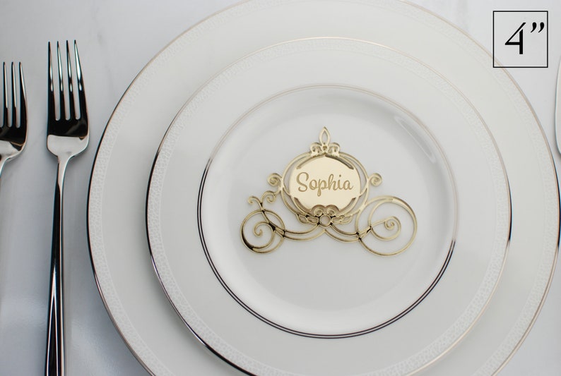 Op de afbeelding: Gouden Assepoester-koets-kaart met de naam Sophia erin geschreven. De kaart is 10 cm hoog en ligt op een wit bord.