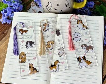 Puede incluir: Cuatro marcapáginas de acrílico transparente con borlas de colores. Cada marcapáginas presenta una ilustración diferente de raza de perro con huellas de patas y corazones. El texto "WOOF!" aparece en dos de los marcapáginas.