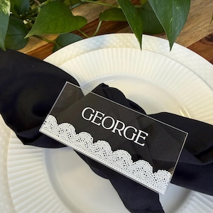May include: A clear acrylic place card with the name "GEORGE" in white, framed by a white lace design. The card rests on a black napkin atop a white plate, with greenery in the background.