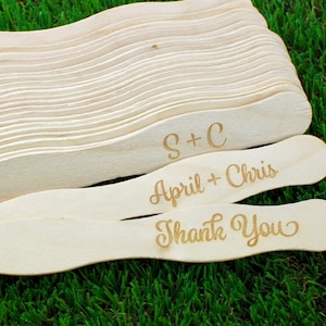 May include: A stack of wooden fans with the text "S+C", "April + Chris", and "Thank You" engraved on them. The fans are on a green surface.